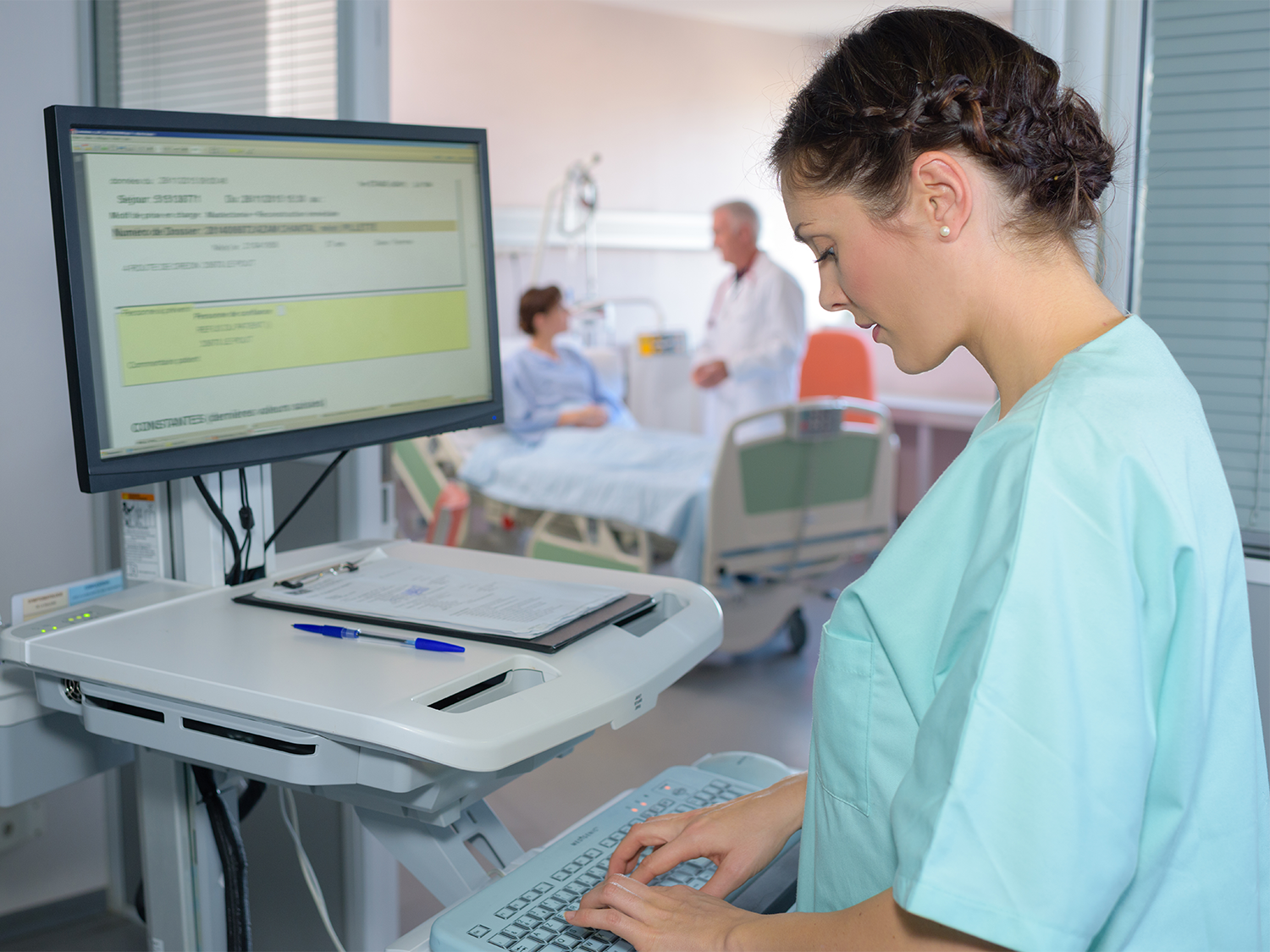 Healthcare professional entering data on a computer with a patient and doctor in the background